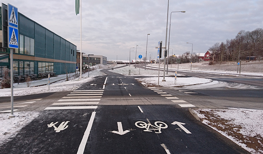 En gång- och cykelväg korsar en bilväg. Till väster står två skyltar som visar att det är ett övergångsställe och en cykelöverfart.