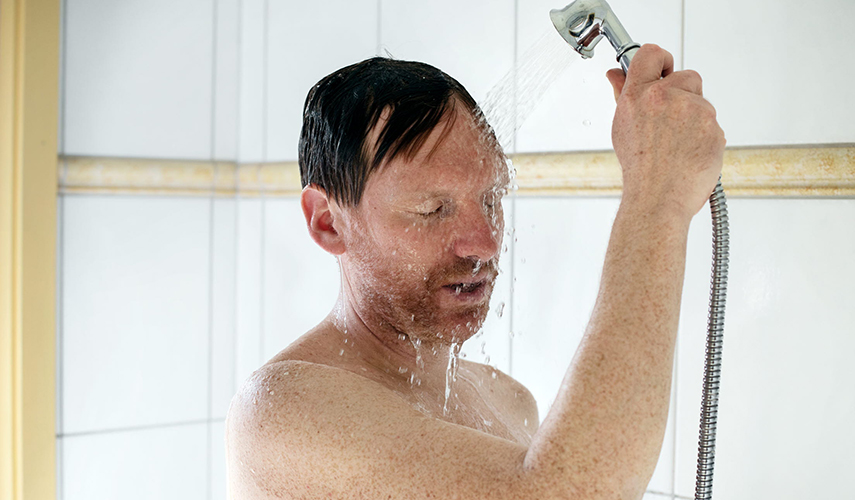 En man står i duschen. Han håller duschmunstycket i högerhanden och vatten rinner ner över hans ansikte.