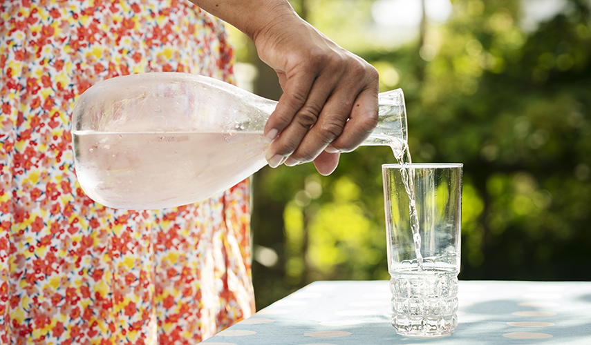 En person i röd blommig klänning håller i en glaskaraff med vatten. Vatten rinner från karaffen ner i ett glas som står på ett vitt bord. i bakgrunden växer gröna buskar.
