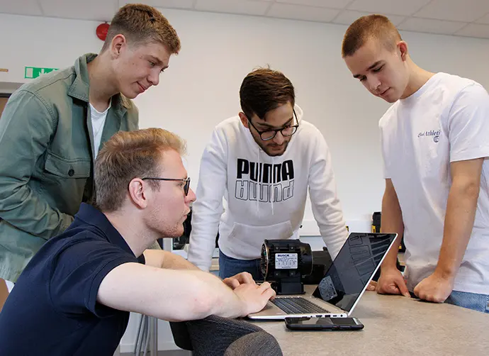 Fyra elever samarbetar kring teknisk utrustning och laptop i klassrumsmiljö.