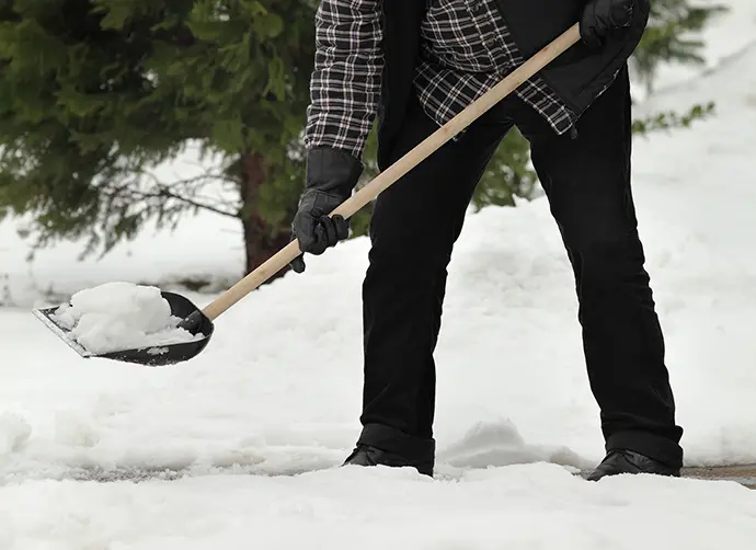En person står på en trottoar och håller i en snöskyffel och skottar snö