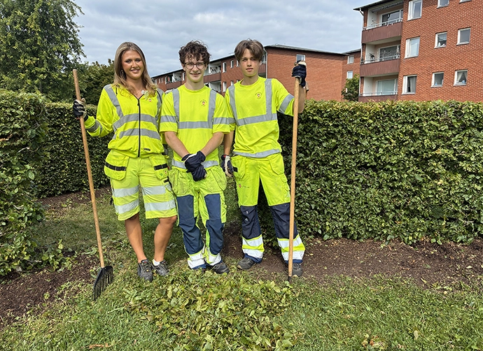 Tre ungdomar i neongula arbetskläder står på gräset framför några häckar. Det finns en hög med löv framför dem. Två av undgomarna håller i ett redskap. De tittar in i kameran. I bakgrunden syns röda tegelhus.