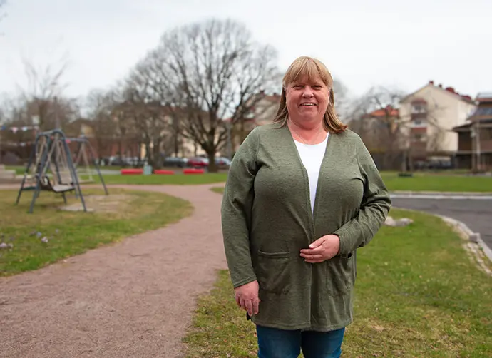 En kvinna står i en park, bärandes en olivgrön cardigan över en vit tröja och jeans. En lekplats syns i bakgrunden.