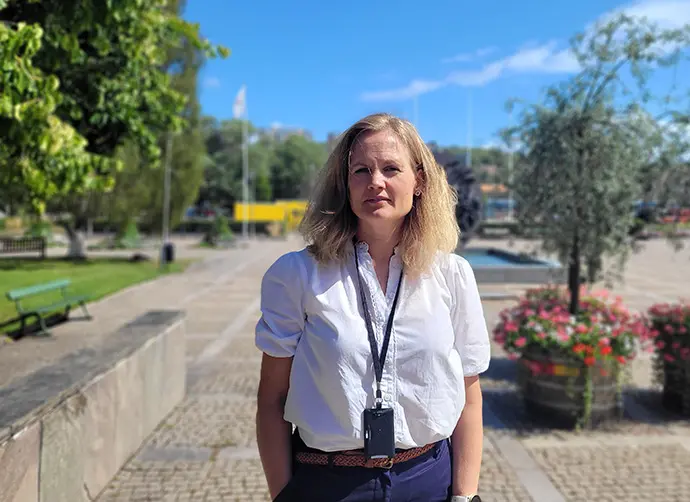 En kvinna står framför stadshusplan där växtlighet syns i bakgrunden. Hon bär en vit blus och mörkblå kjol.