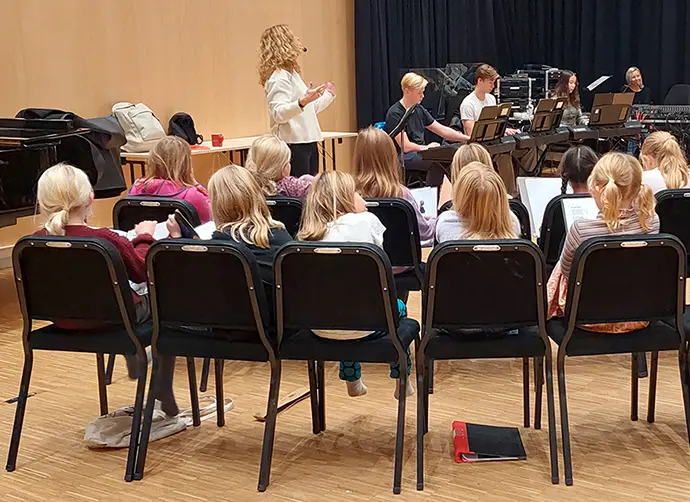 En grupp barn sitter med textblad på stolar och sjunger framför en lärare med headset-mikrofon. Fyra ungdomar spelar keyboard i bakgrunden.