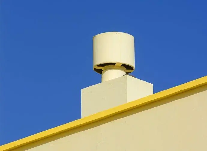 En vit ventilationskanal på en ljus byggnad med gult tak mot en blå himmel.