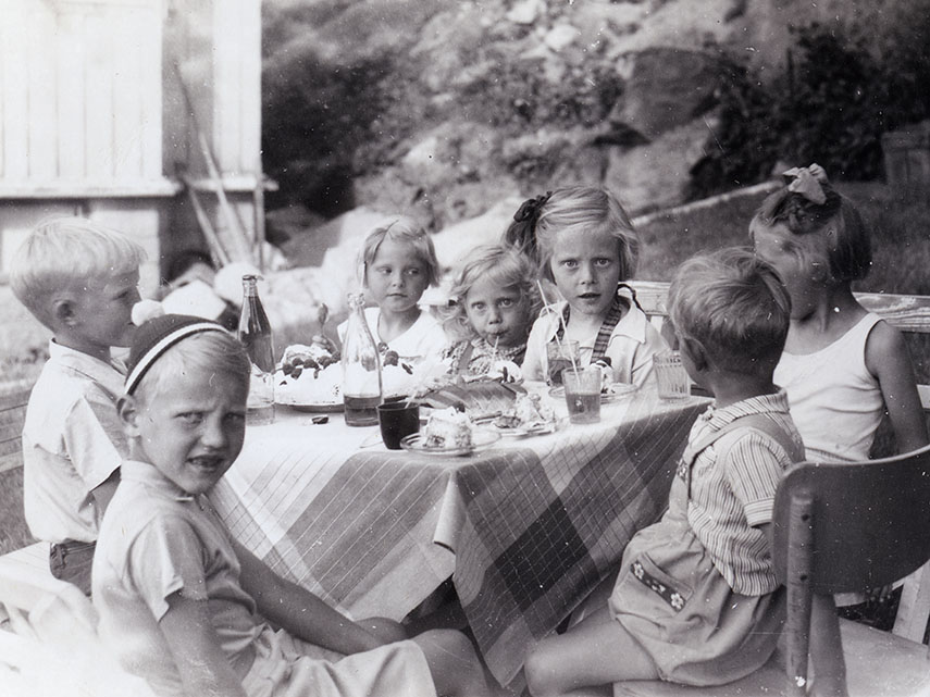 Ett svartvitt foto. En samling barn sitter runt ett bord med fika utomhus.