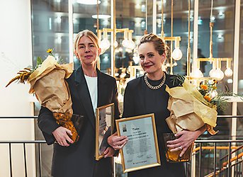 Två kvinnor står och håller varsin blombukett och varsitt diplom i ram.