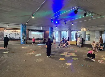En mängd människor är samlade på en asfalterad yta under ett betongtak. De målar, leker och i bakgrunden visas ett bildspel.